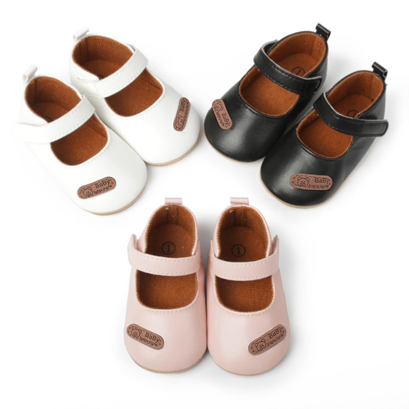 Three pairs of leather baby Mary Jane shoes in white, black, and pink with Velcro straps and brown patches.