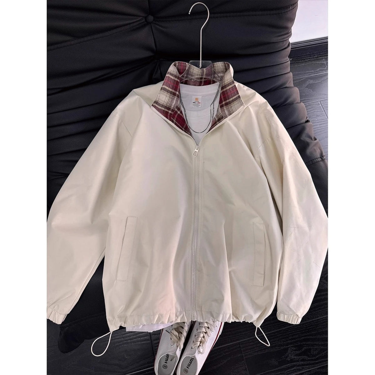 Off-white zip-up jacket with a red plaid collar, drawstring hem, and side pockets, hanging on a hanger.