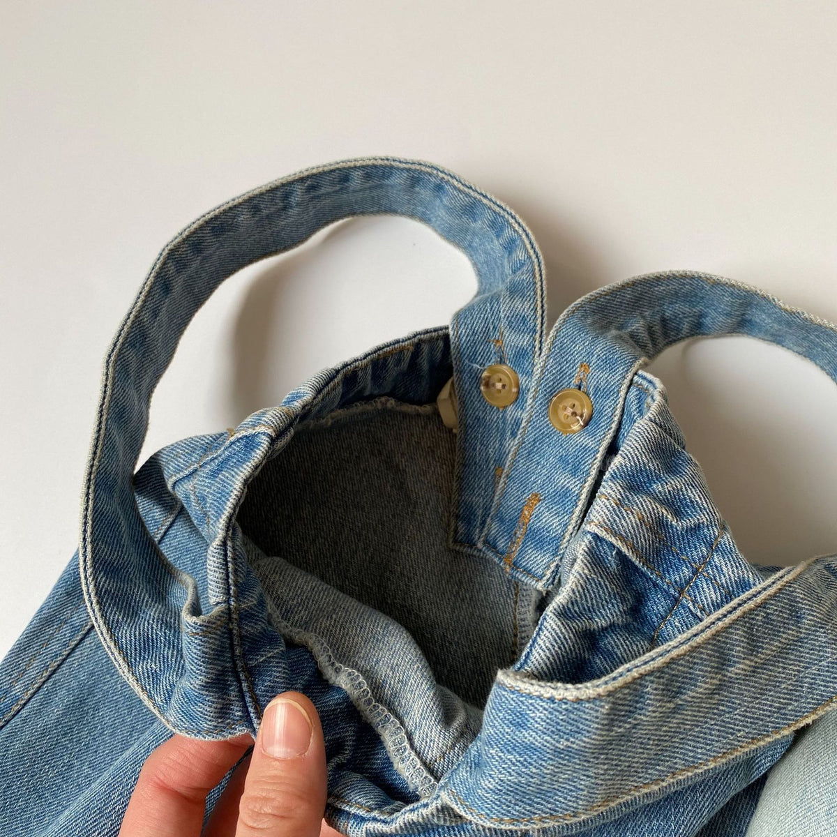 Blue denim tote bag with brown buttons and shoulder straps, held by a hand.