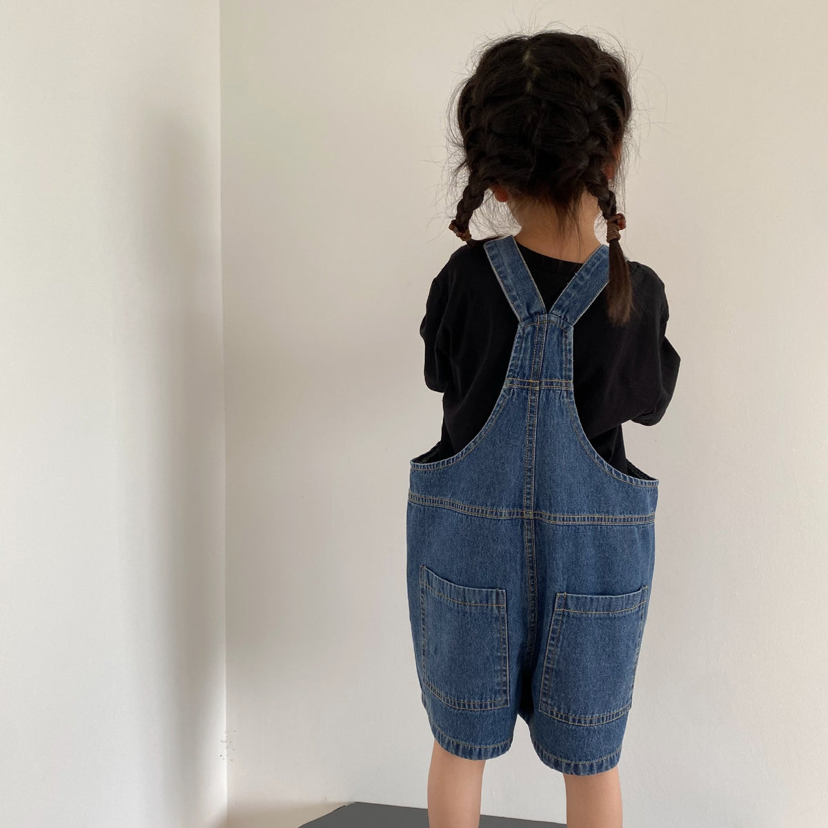 Child seen from behind wearing blue denim overalls and a black shirt against a plain wall.