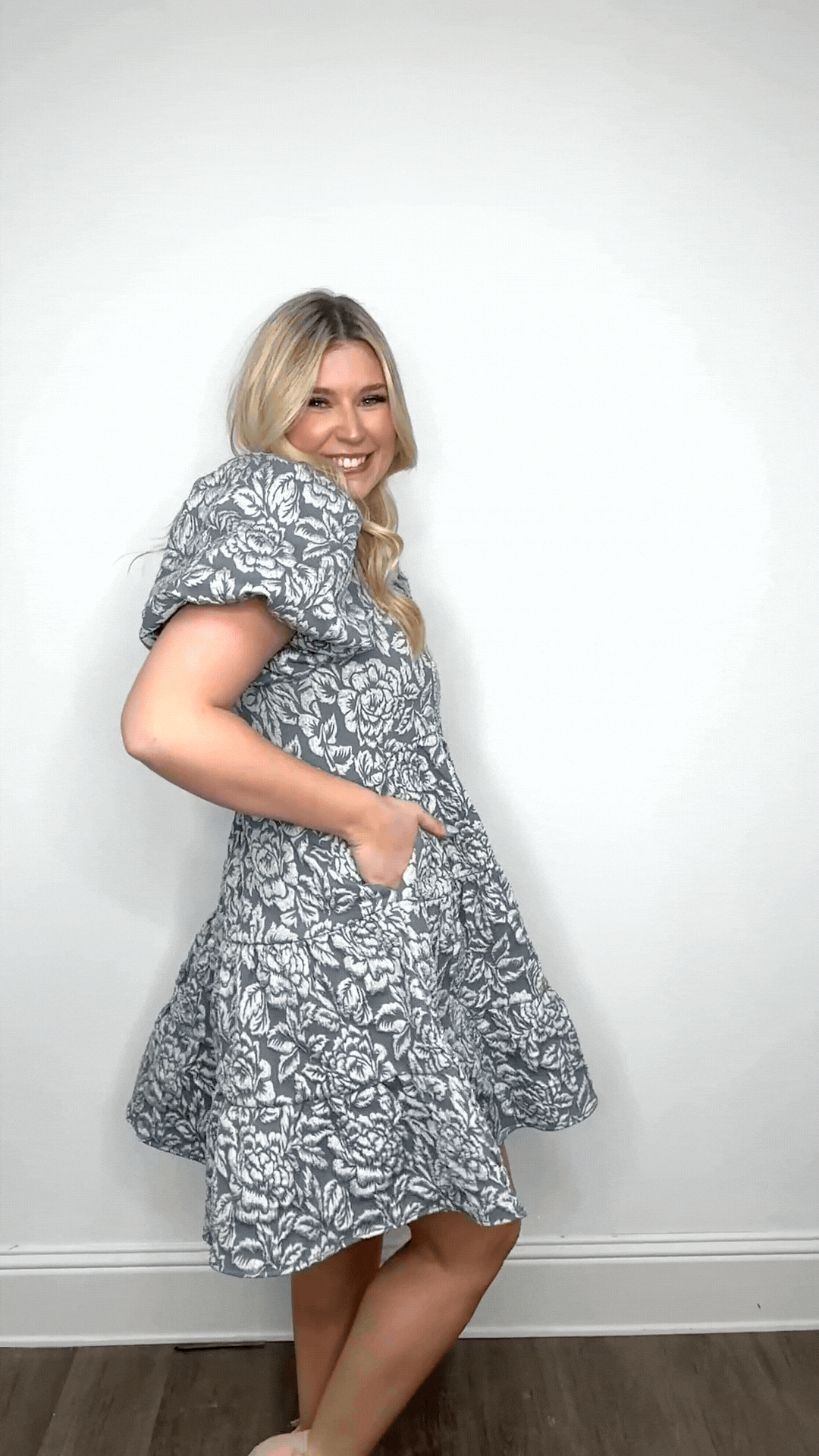 A smiling blonde woman models a blue and white floral jacquard short dress with puffed sleeves and a tiered skirt.