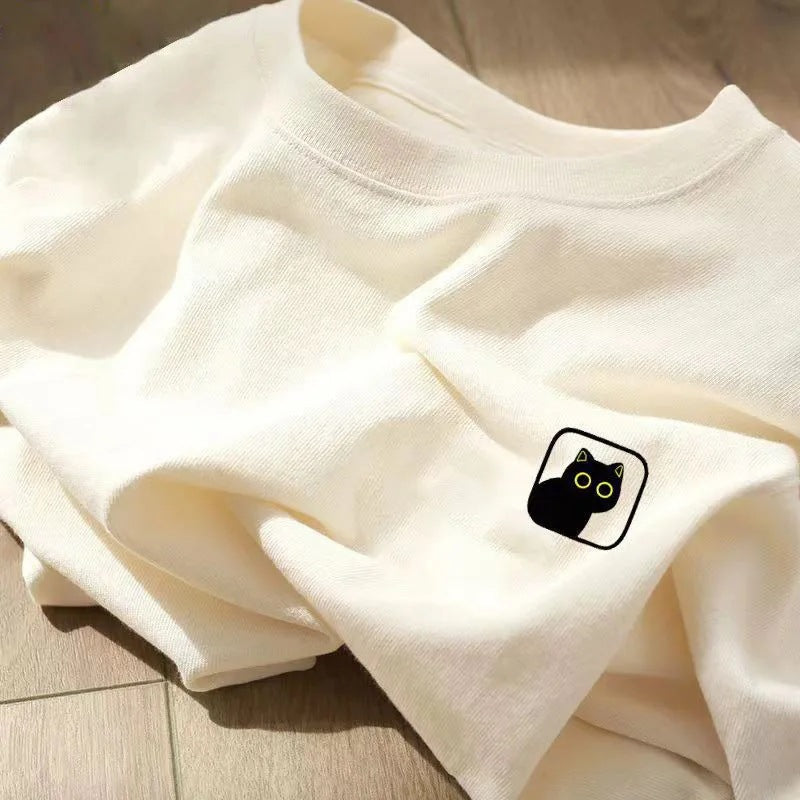 Folded off-white round-neck t-shirt featuring a small black cat patch with yellow eyes.