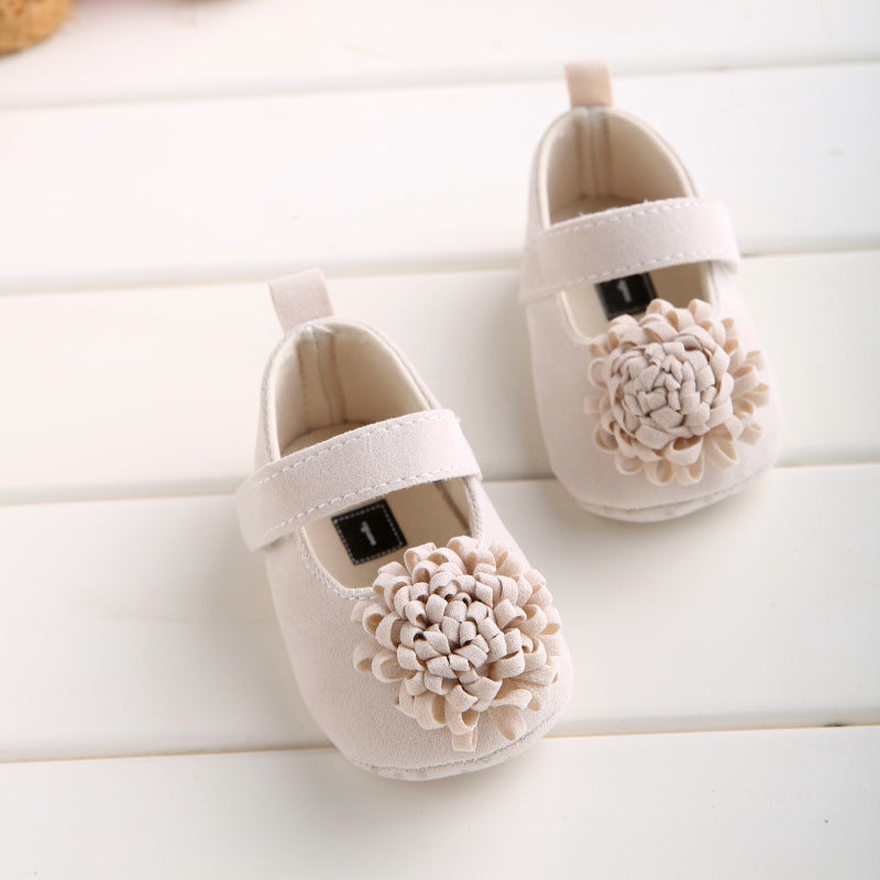 Beige suede baby shoes with velcro straps, feature a large fabric flower and a small tag with the number 1 inside.