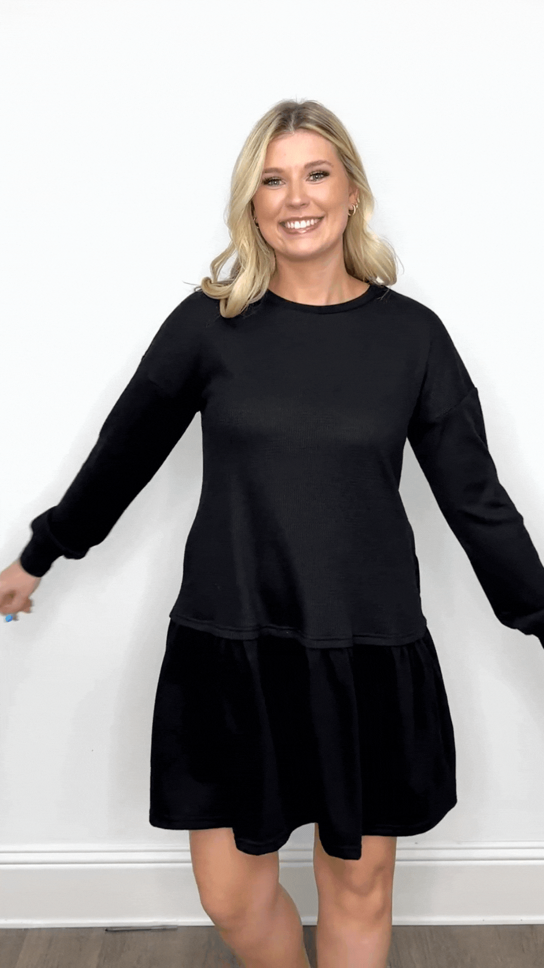 A smiling blonde woman models a black waffle knit long-sleeve dress with a dropped waist and flared skirt against a plain white wall.