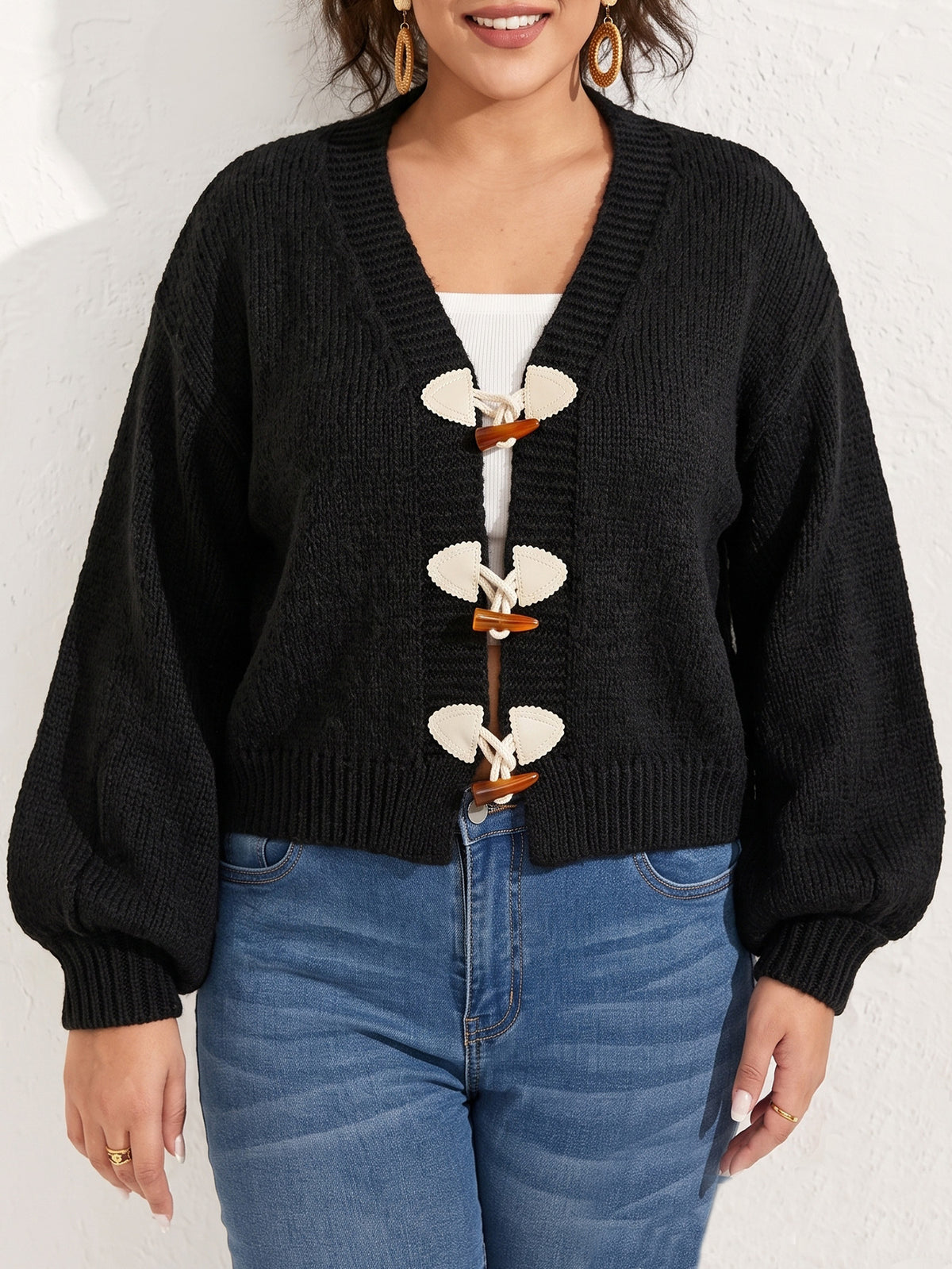 A plus-size woman wearing a black knit cardigan with three toggle buttons over blue jeans.