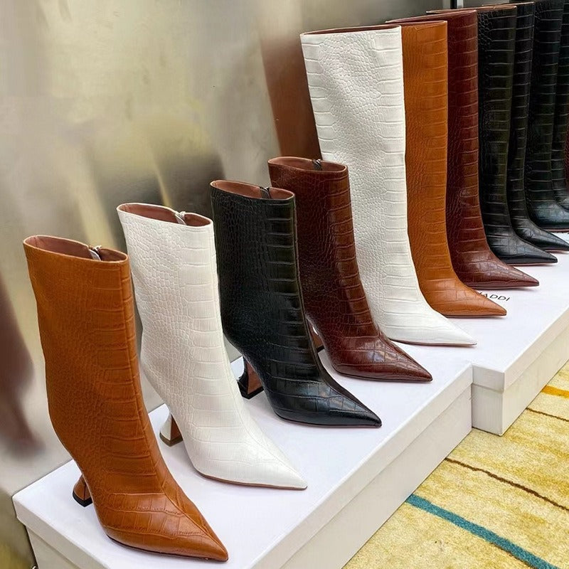 A row of pointed-toe crocodile-embossed leather boots in tan, white, black, and brown, featuring flared heels.