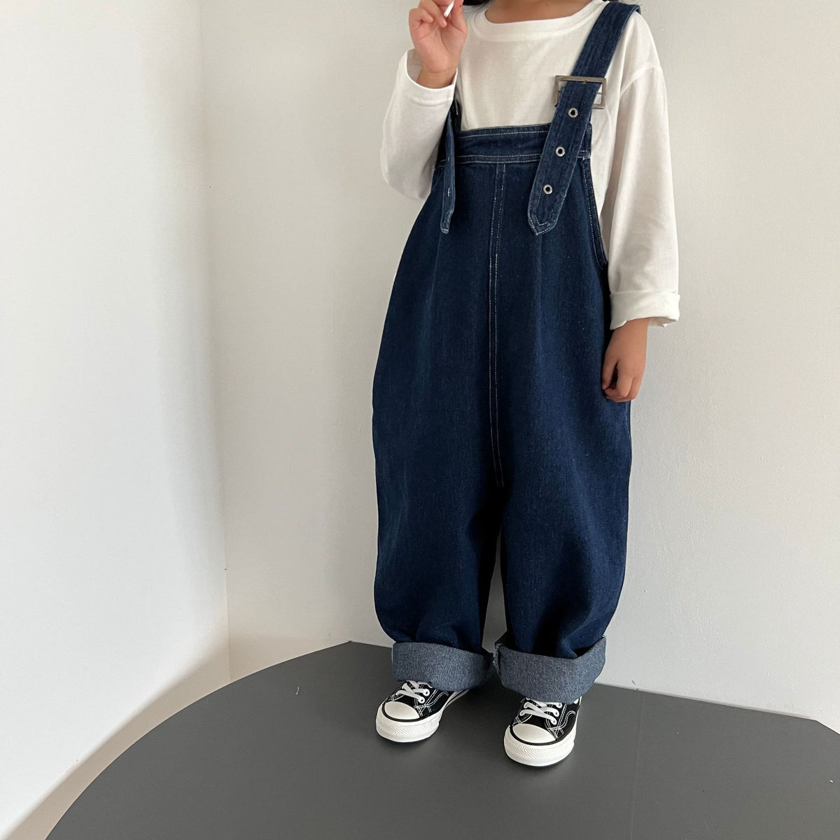 Dark blue denim overalls with cuffed hems and button straps over a white long-sleeve shirt.