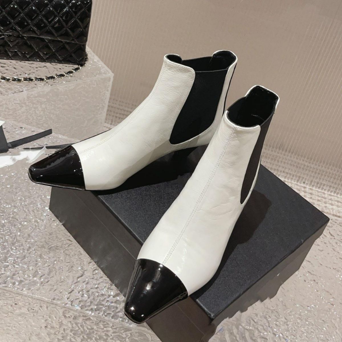 White leather ankle boots with black patent toes and side panels, pointed toes, and low heels on a box.