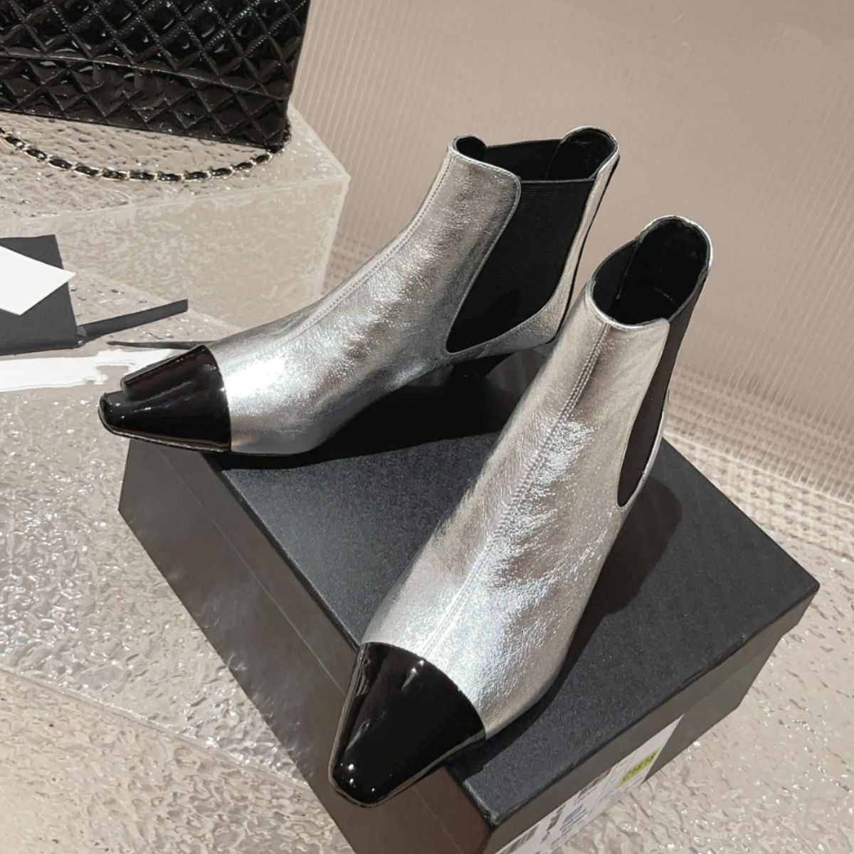 Silver and black leather Chelsea boots with contrasting cap toes, elastic side panels, and low heels on a box.