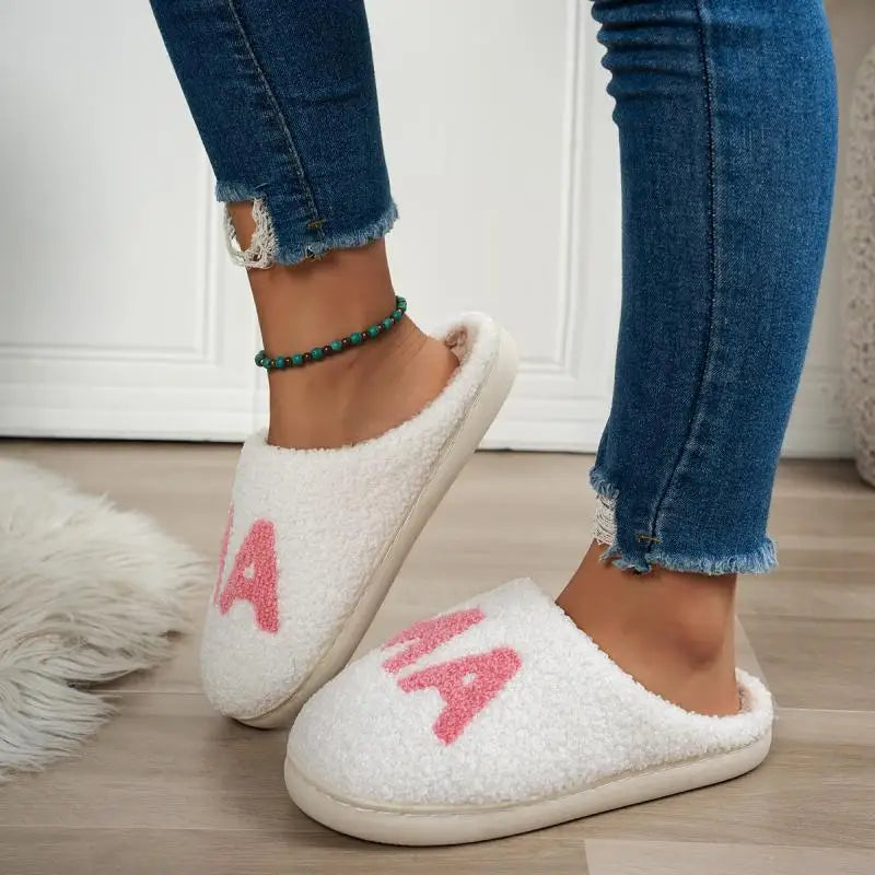 White fuzzy slip-on slippers with pink chenille lettering "MAMA" on the top, worn with blue jeans.