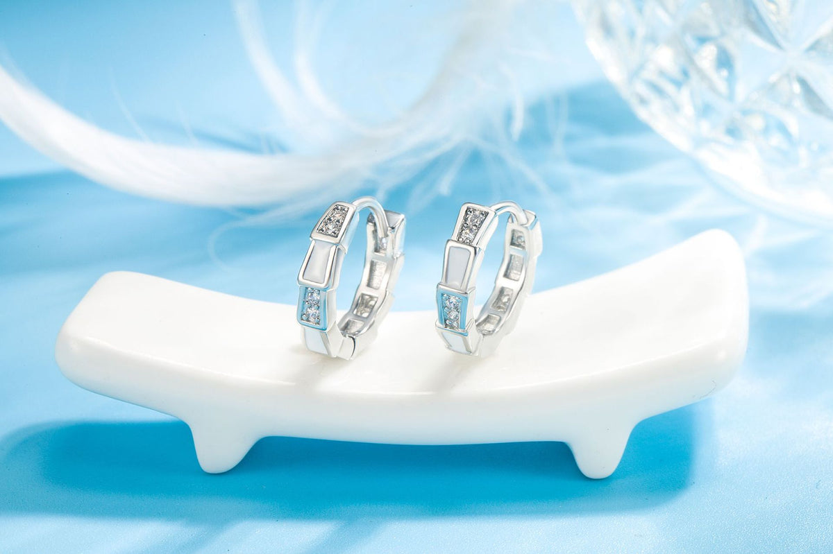 Silver hoop earrings with white enamel links and embedded crystals on a white ceramic stand.