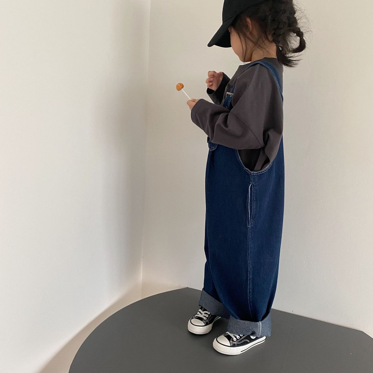 Child in dark denim overalls, grey long-sleeve shirt, and black sneakers, holding a lollipop.