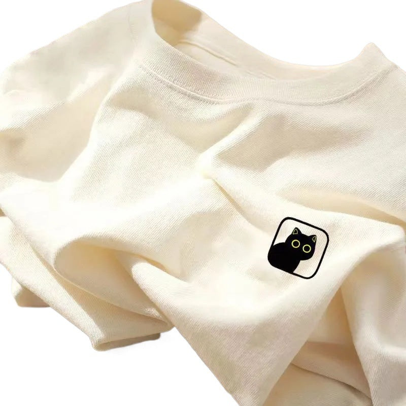 A folded cream round-neck T-shirt features a small black cat graphic patch on the chest.