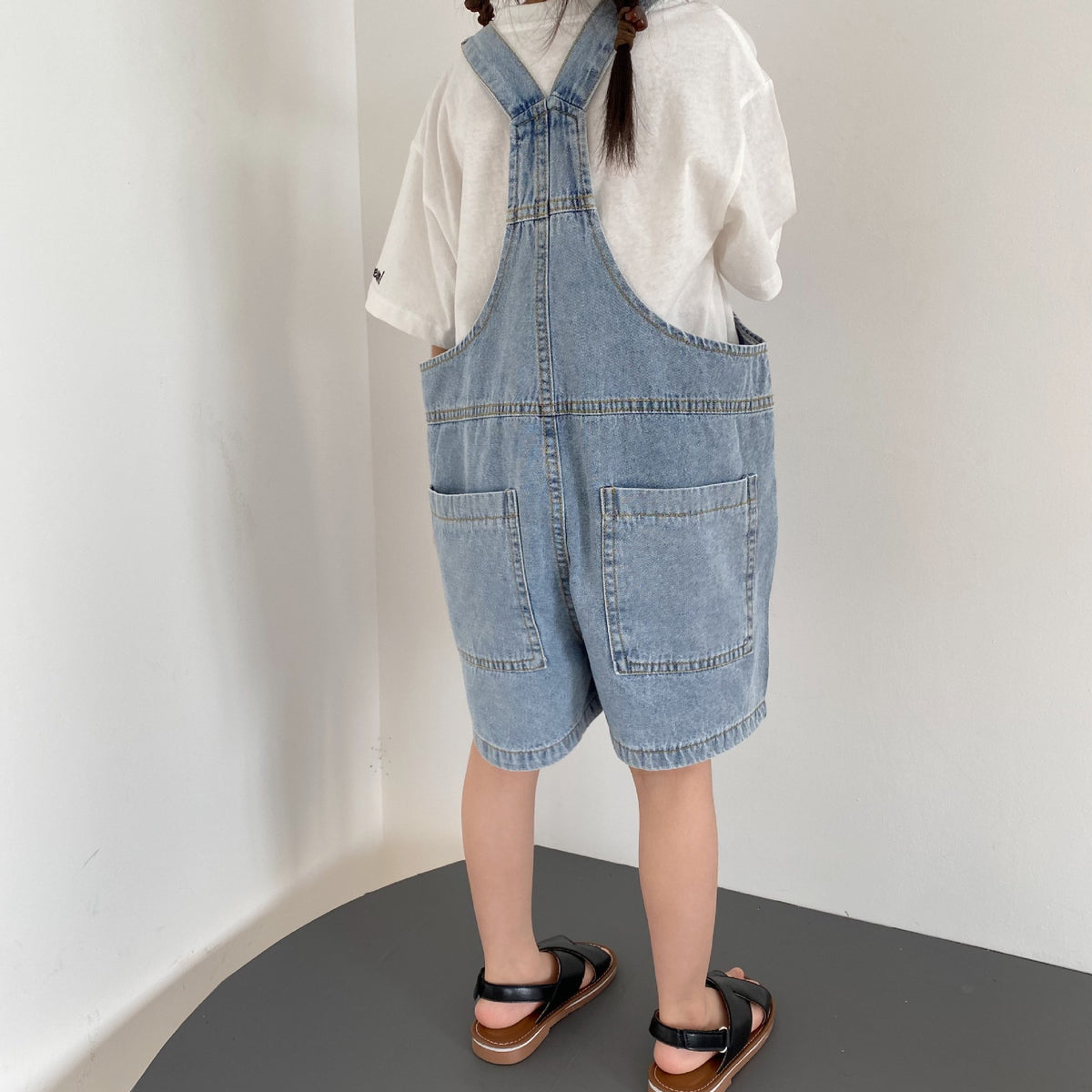 Child wearing blue denim overalls, a white t-shirt, and black sandals seen from behind.