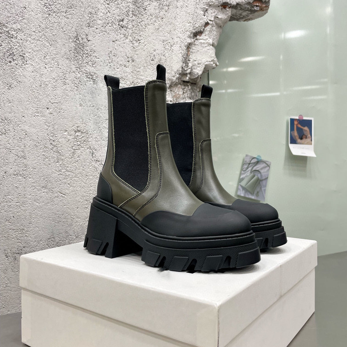 Olive and black leather Chelsea boots with chunky lug soles and pull tabs on white box.
