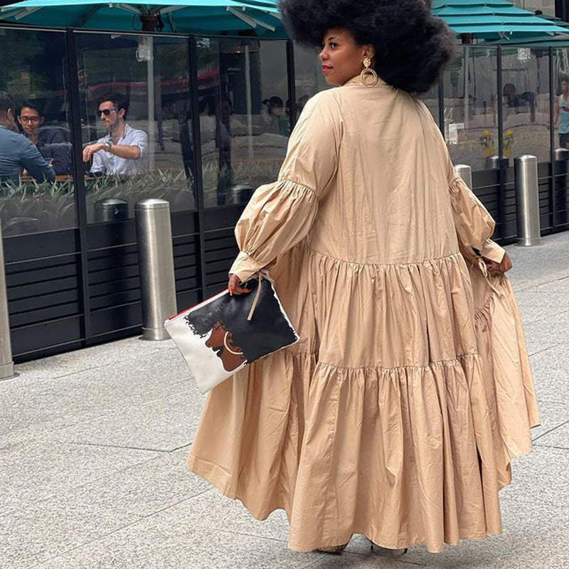 A woman wearing a beige cotton maxi dress with long tiered ruffles, balloon sleeves, and a matching clutch.