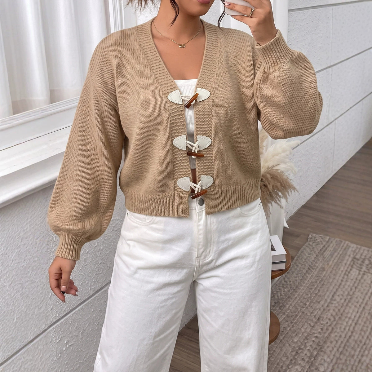 A woman wearing a tan knitted short cardigan with horn toggle buttons over a white top and pants.