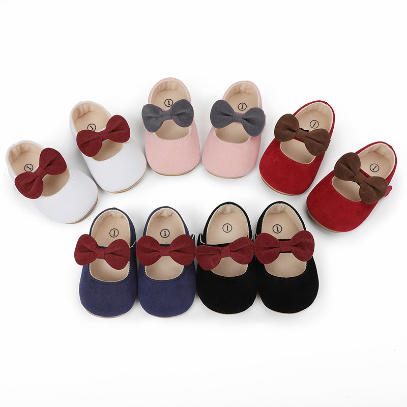 Six pairs of suede baby crib shoes with bow accents in white, pink, red, navy blue, and black.