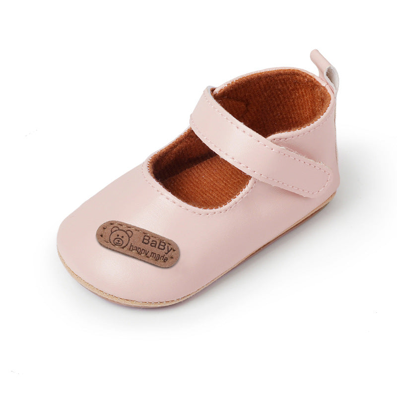 Pink leather baby shoe, Mary Jane style with hook-and-loop strap, brown interior, and branded patch.
