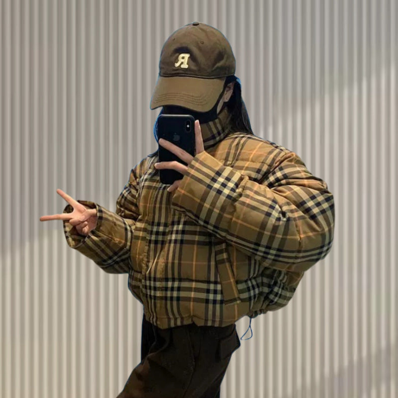 A woman in a plaid puffer jacket and brown cap poses, holding a phone in a mirror.