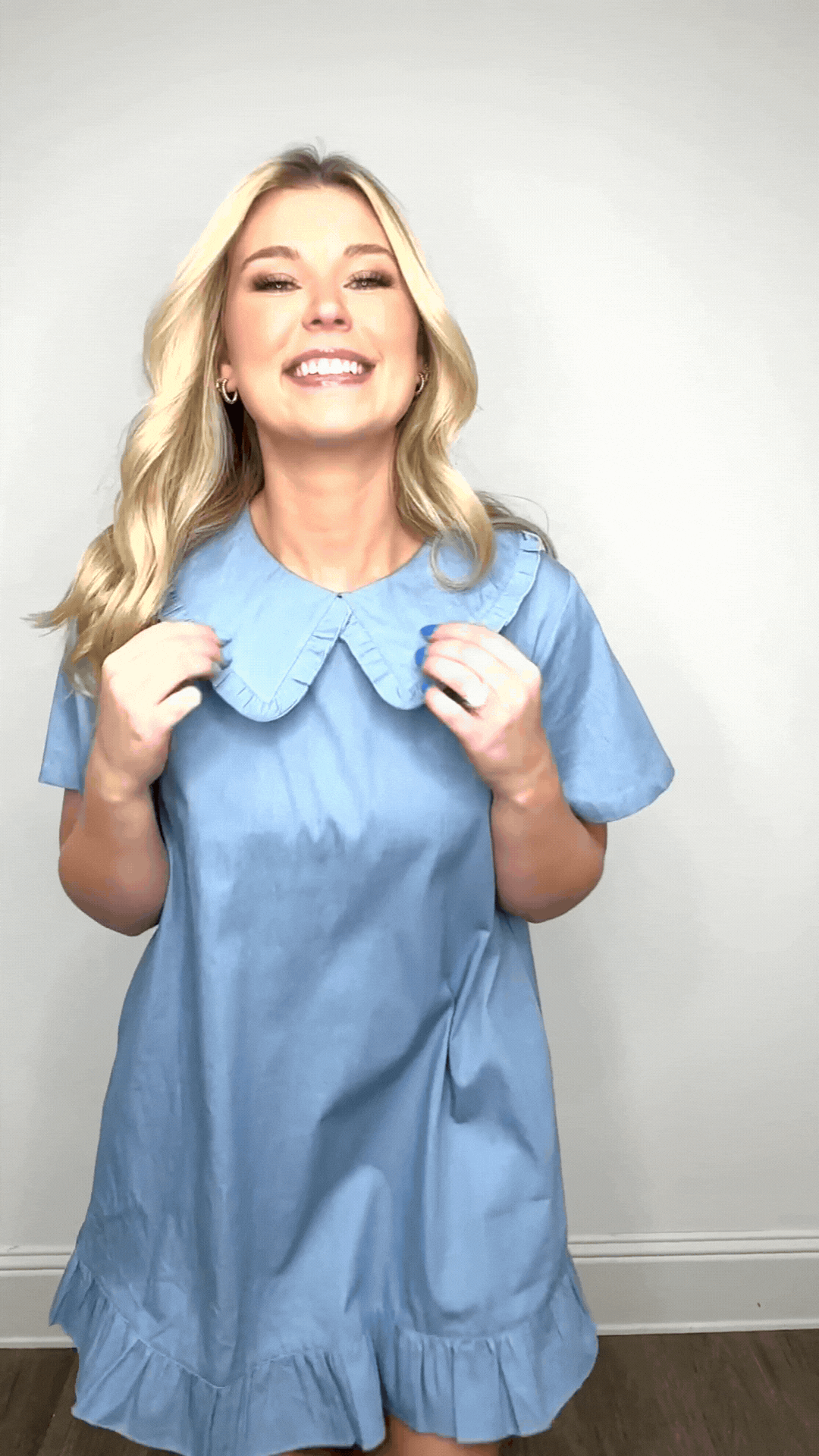 Smiling blonde woman holding the collar of a light blue chambray dress with ruffles.