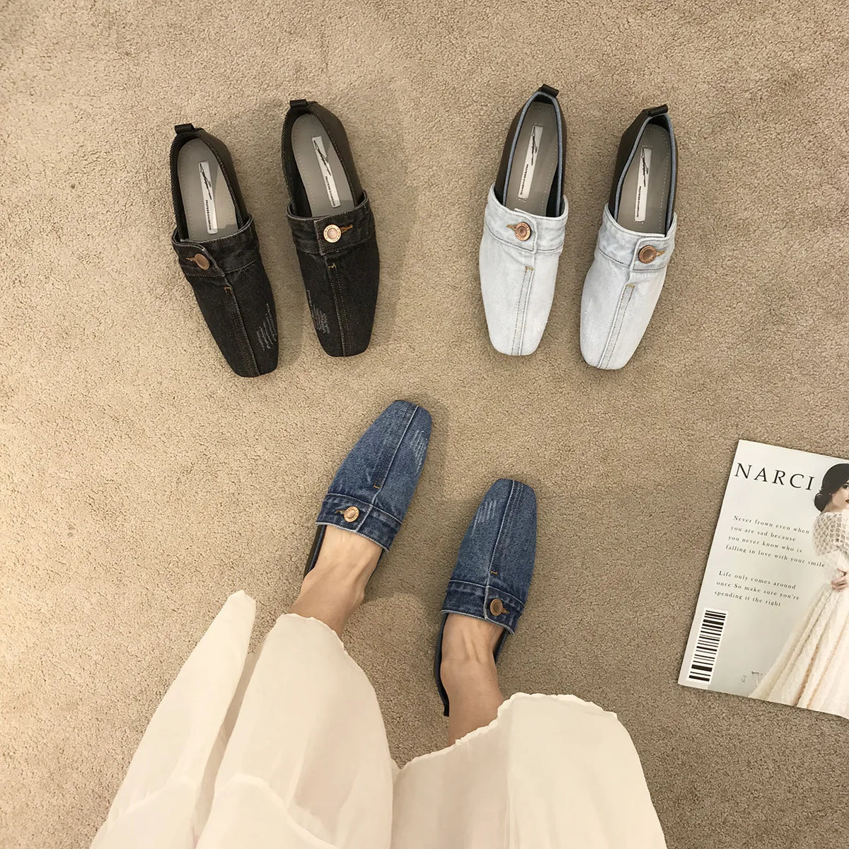 Blue denim slip-on square-toe loafers with gold button accents on the vamp, arranged on a textured beige surface.