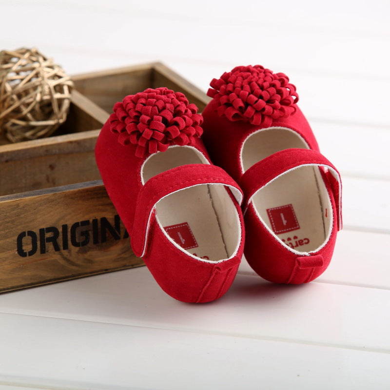 Red suede baby shoes with a floral pom-pom and hook-and-loop strap, beside a wooden box.