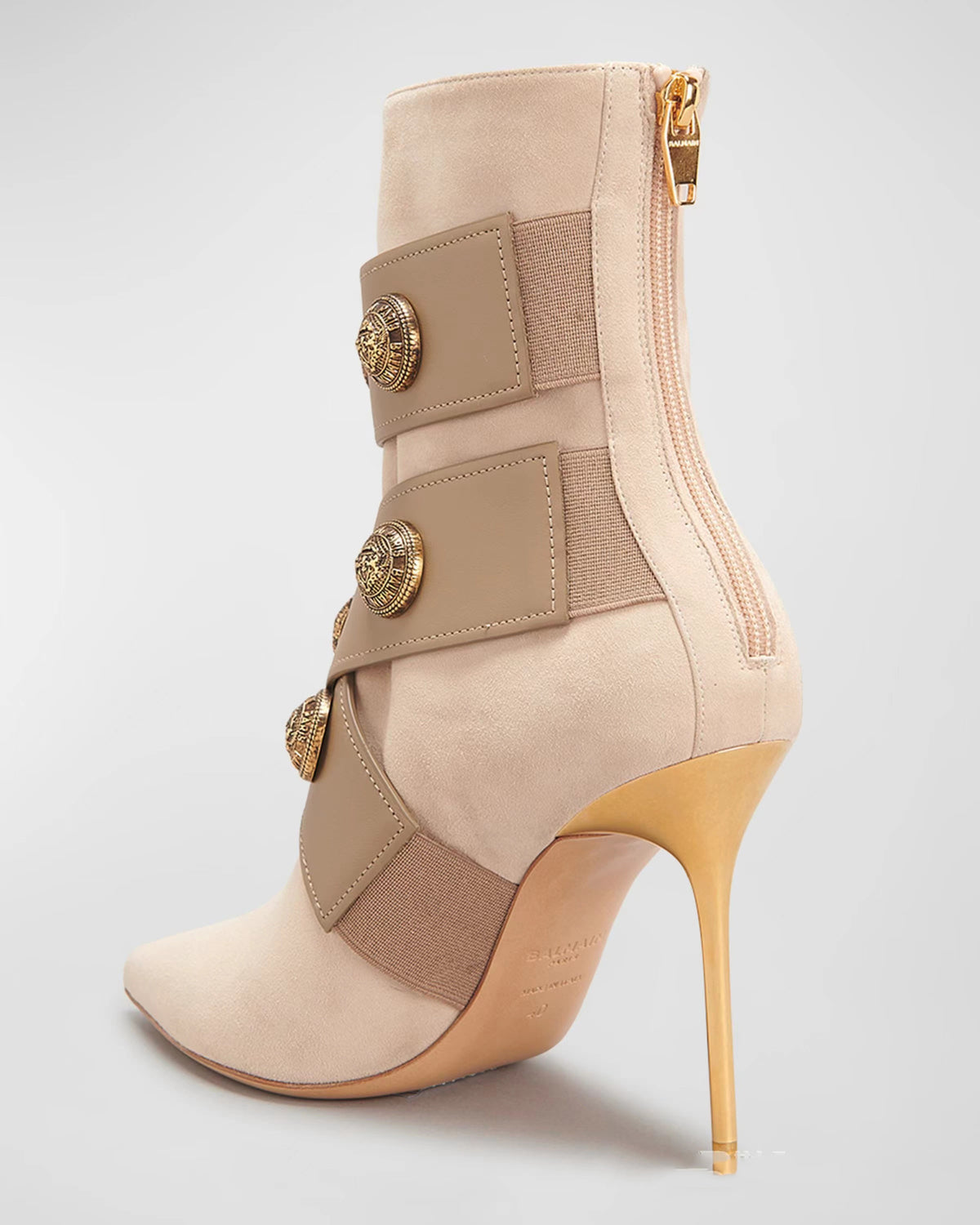 Beige suede stiletto boot with gold heel, back zipper, and buckled straps featuring large embossed buttons.