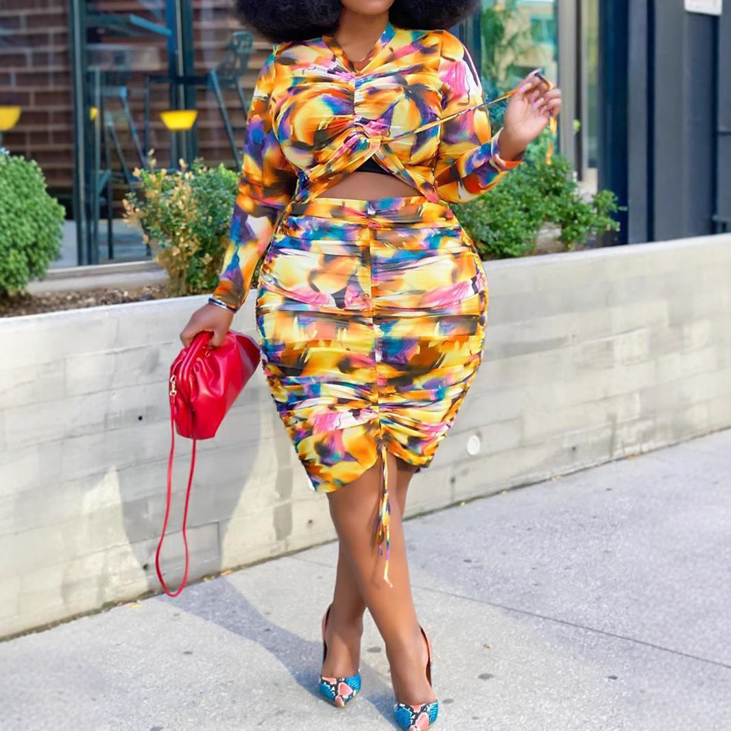Colorful abstract two-piece set with long-sleeve crop top and ruched midi skirt, holding a red clutch.
