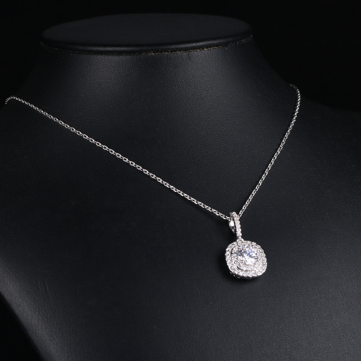Silver metal chain necklace with a round pendant surrounded by small sparkling stones on a black display.
