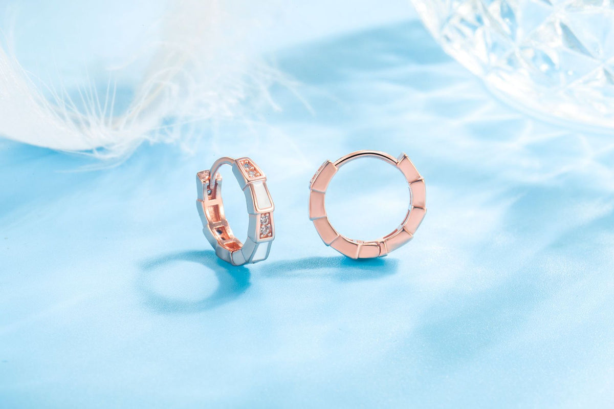 Rose gold huggie hoop earrings featuring segmented geometric patterns with white enamel and sparkling stone accents.