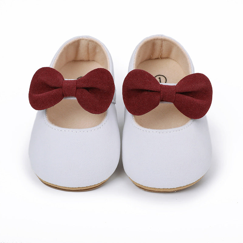 White baby shoes with a maroon suede bow across the front, featuring a classic round-toe Mary Jane style.