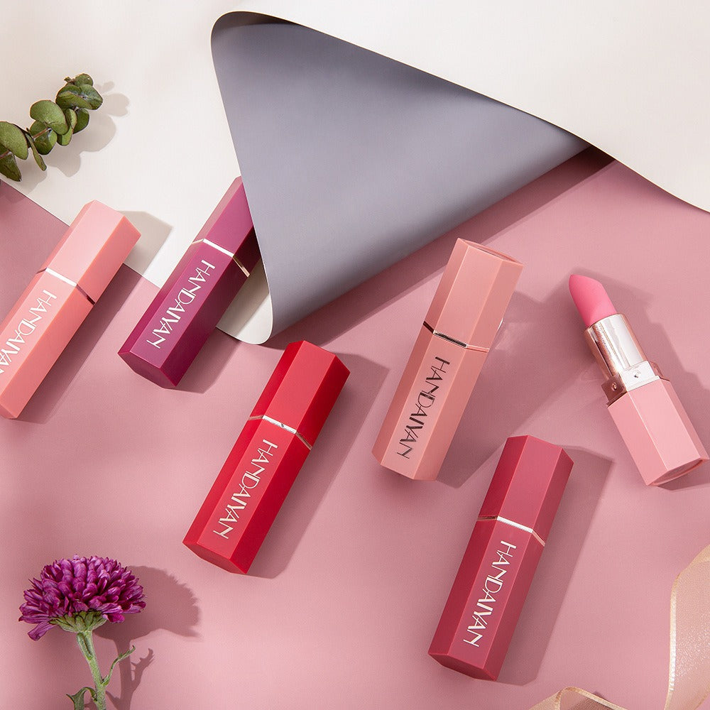 Six pink hexagonal plastic lipsticks labeled "HANDAIYAN" lie on a pink and gray backdrop with a flower.