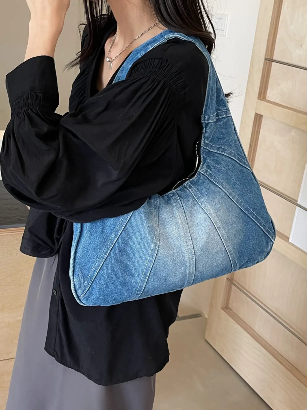 Blue denim slouchy shoulder bag with paneled seams and a curved zip top, worn over a black top.