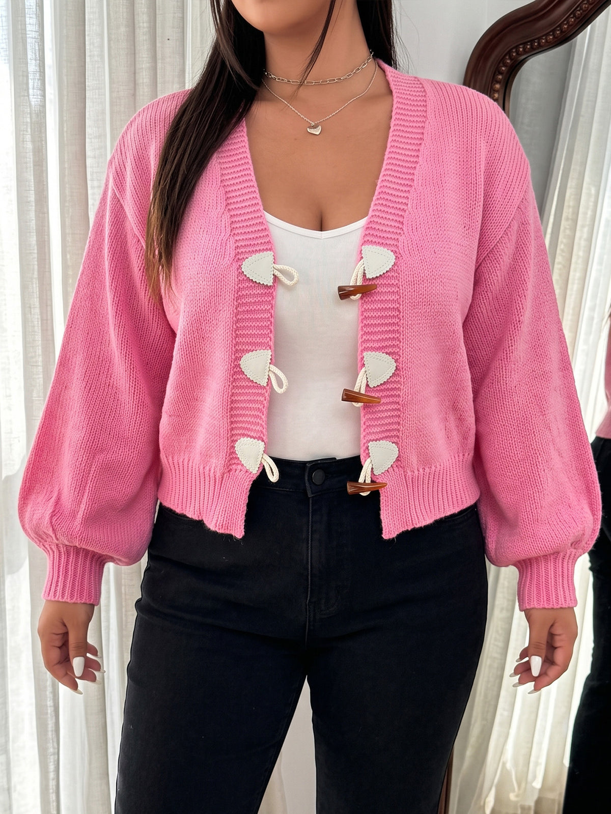 A woman wears a pink knit cardigan with toggle buttons and long sleeves, over a white top and black jeans.