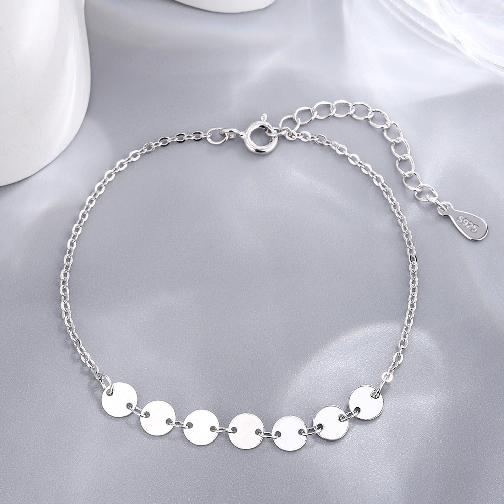 A silver chain bracelet featuring a row of small, flat round discs on a grey cloth.