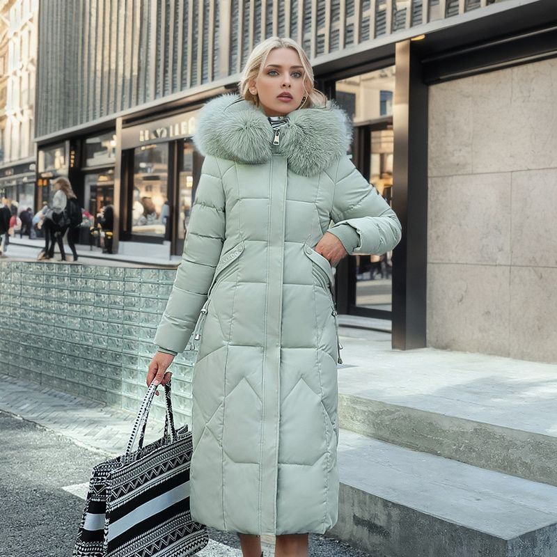 A woman wearing a knee-length, quilted sage green puffer coat with a large faux-fur hood.