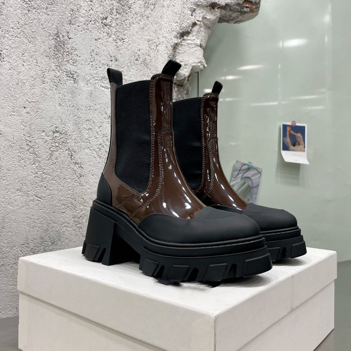 Brown patent leather Chelsea boots with black rubber lug soles and toe caps on a white box.