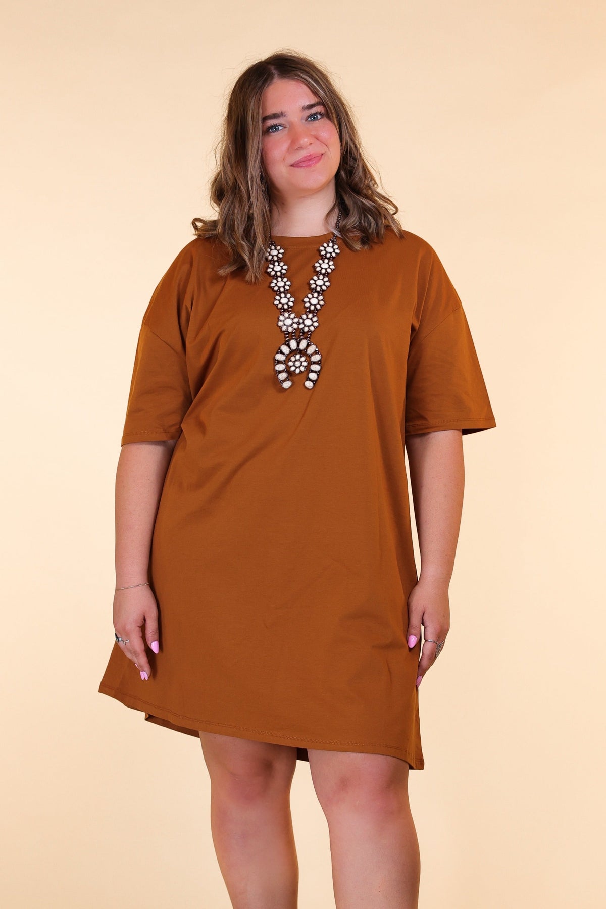 A woman poses in a solid rust t-shirt dress with short sleeves, accessorized with a large statement necklace, against a plain beige background.