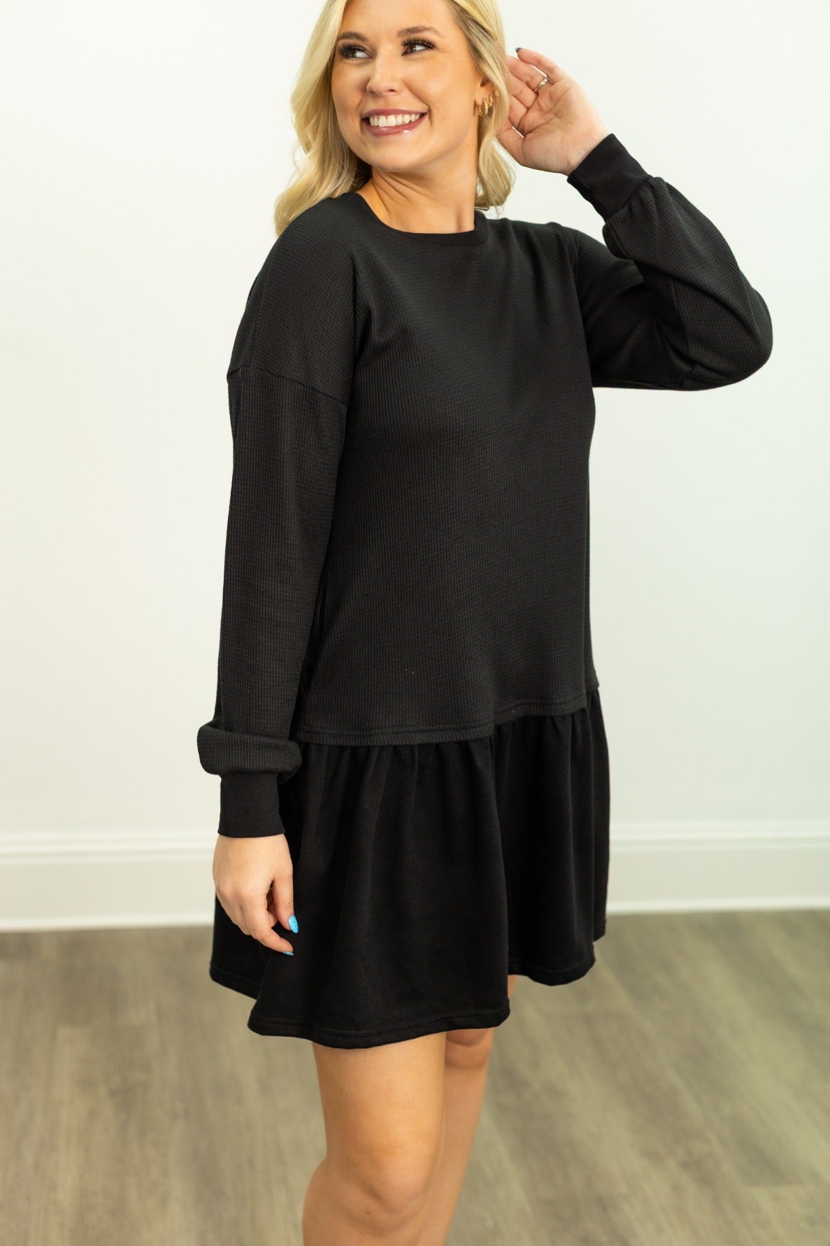 Blonde woman wearing a black waffle-knit long-sleeve dress with a ruffled hem, posing indoors.