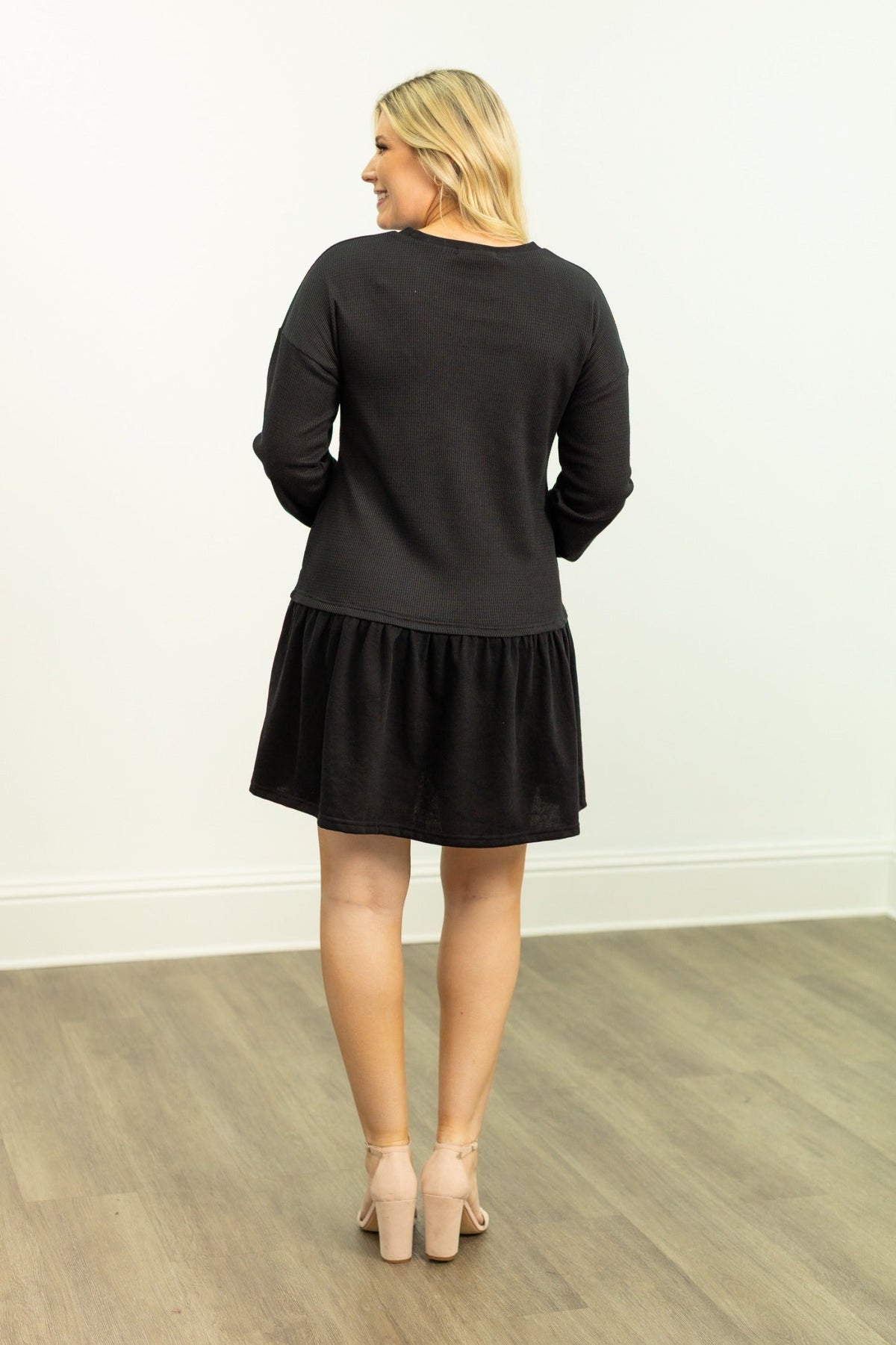 Back view of a blonde woman wearing a black waffle knit long-sleeve dress and heeled sandals against a white wall.