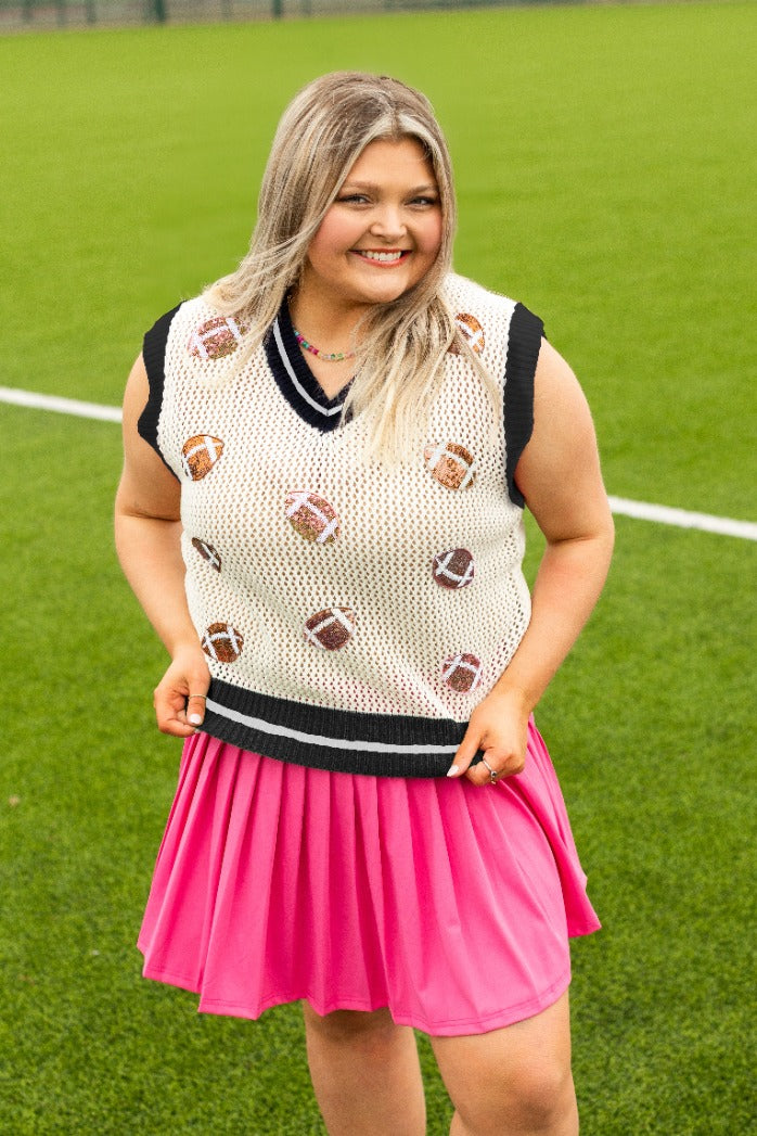 A smiling blonde woman in a white knit football patch vest and pink pleated skirt on turf.