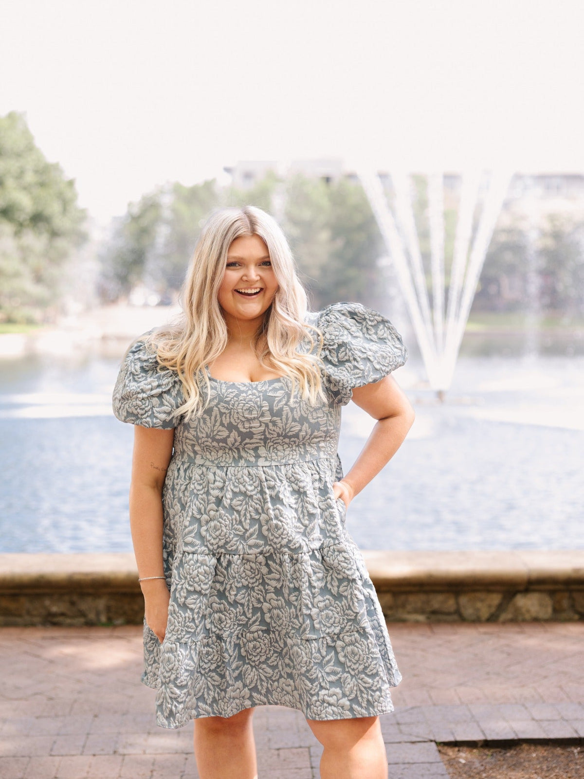 Blonde woman wearing a blue jacquard floral mini dress with puff sleeves, posing near an outdoor fountain.