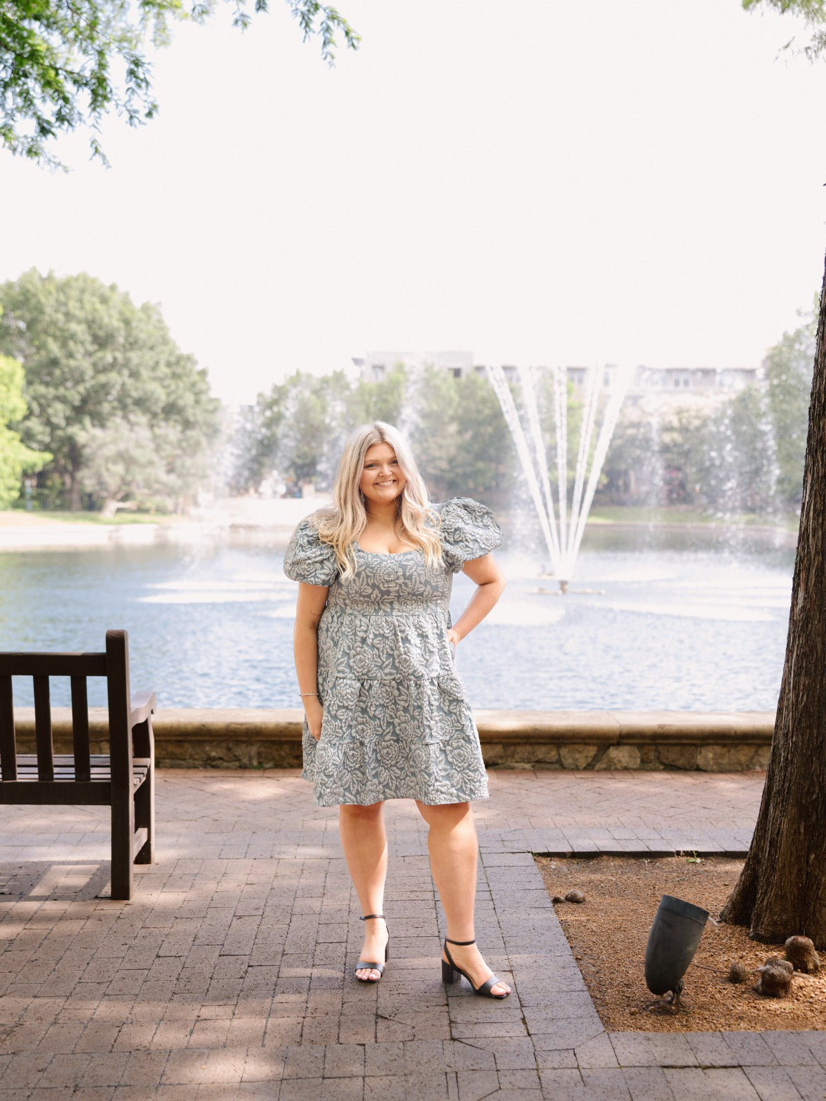 Blonde woman wearing a blue jacquard tiered dress with puff sleeves, standing on a brick path by a fountain.