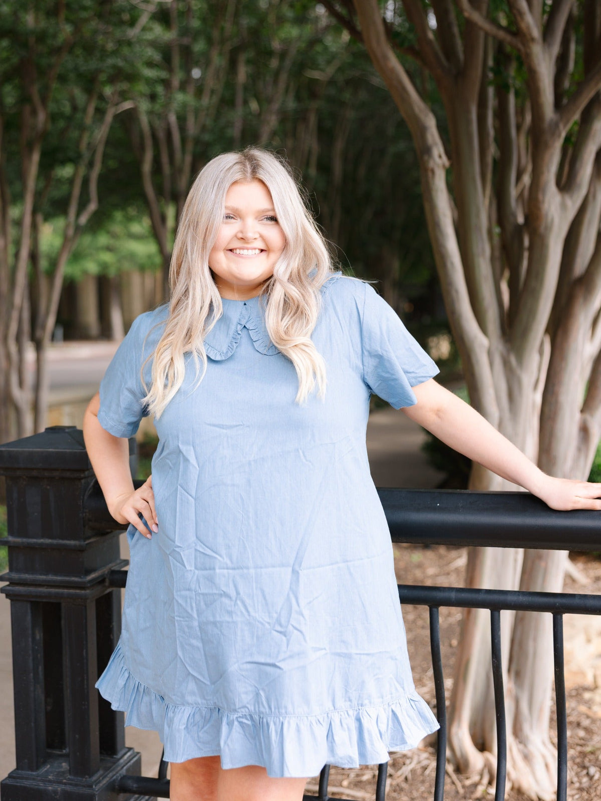 Blonde woman wearing a light blue chambray dress with a wide ruffled collar and hem, posing outdoors by a railing.