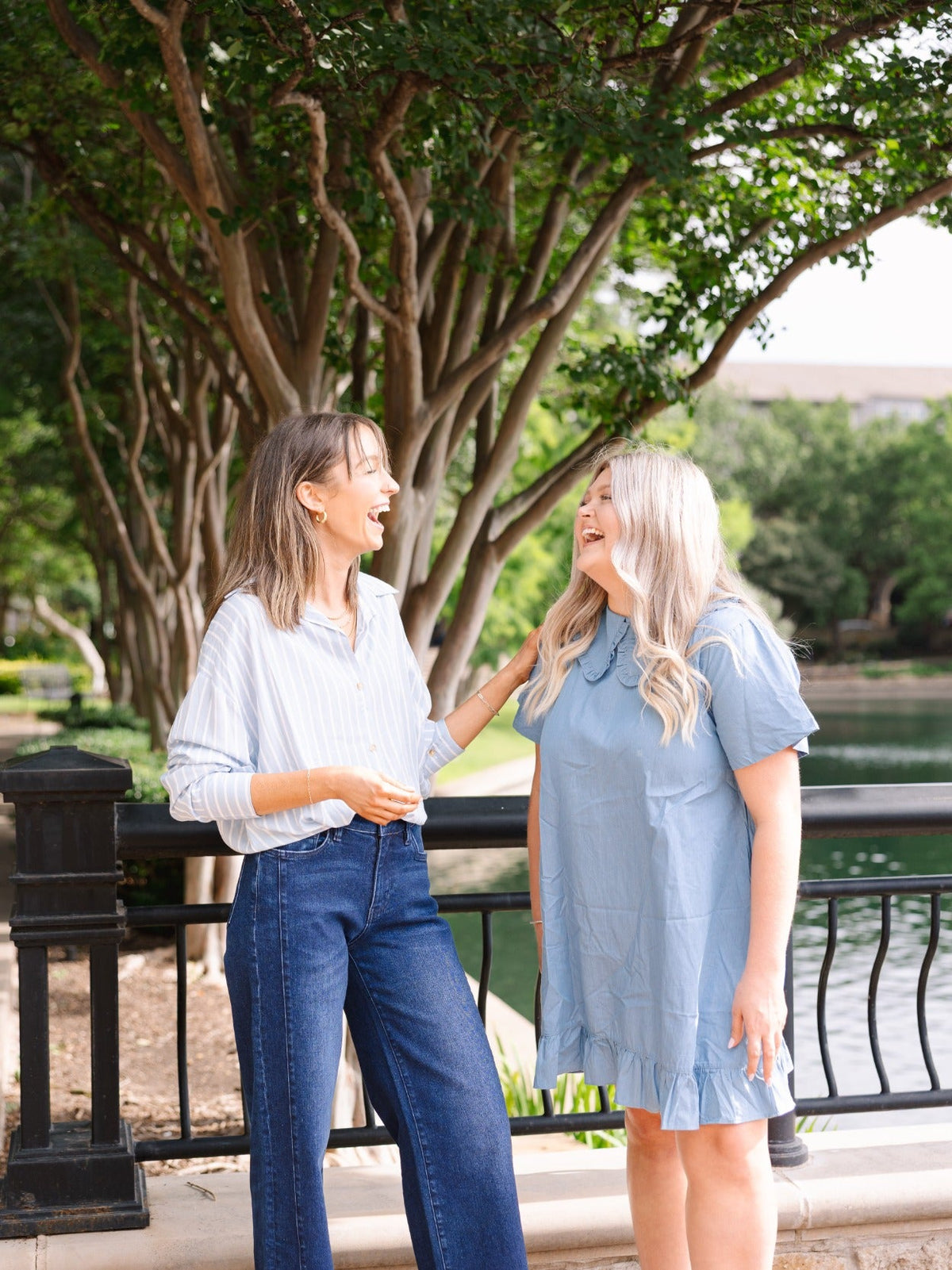 Two women laughing outdoors near a railing; one wears a blue chambray dress with a wide collar and ruffled hem.