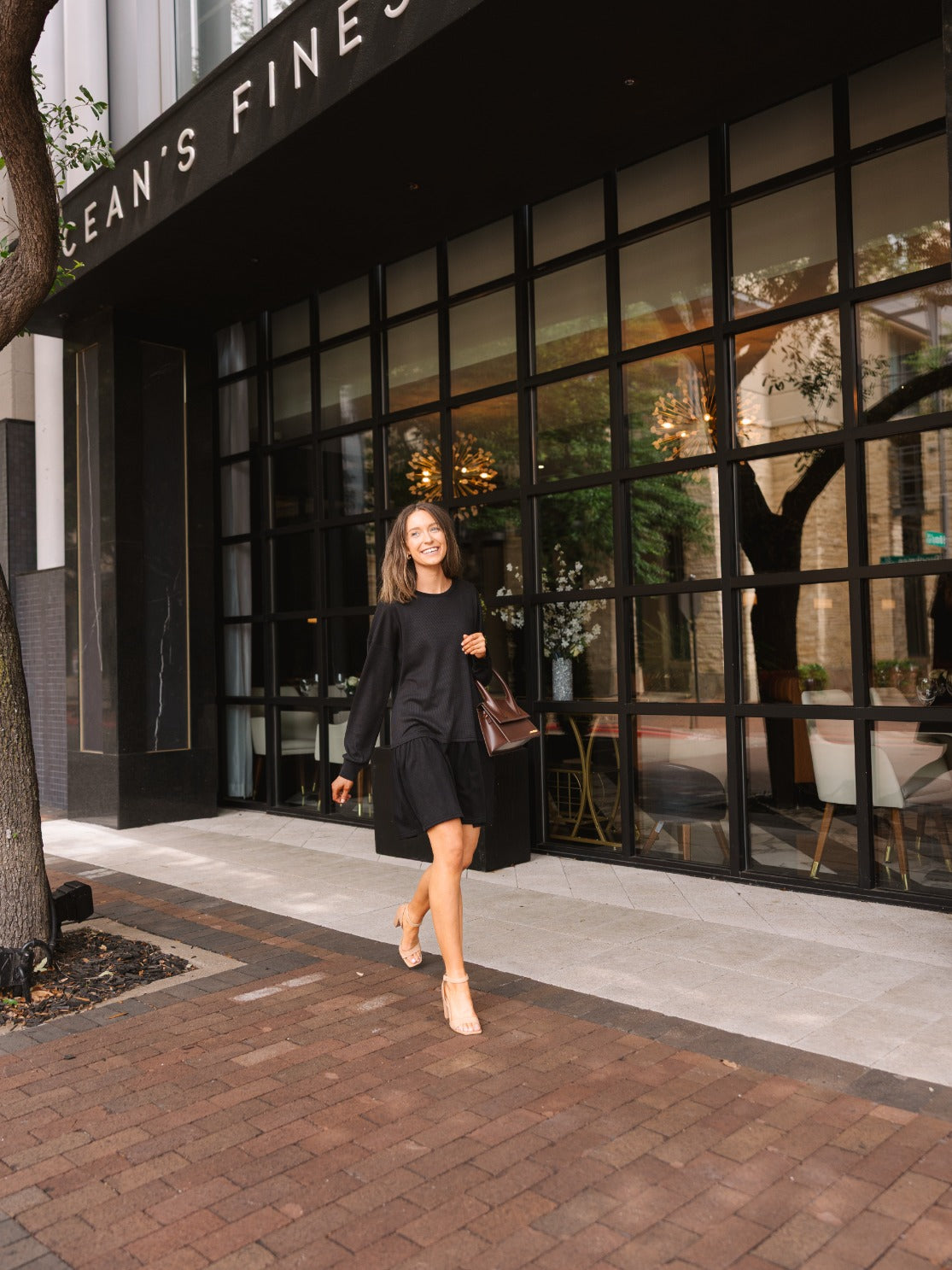 A smiling woman walks on a sidewalk in a black long-sleeve waffle-knit dress, carrying a brown handbag.