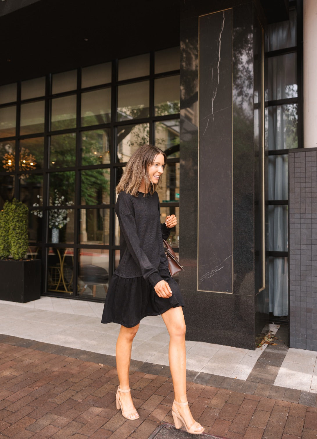A woman walking outdoors wears a black waffle-knit long-sleeve dress with a drop waist, paired with nude heels.