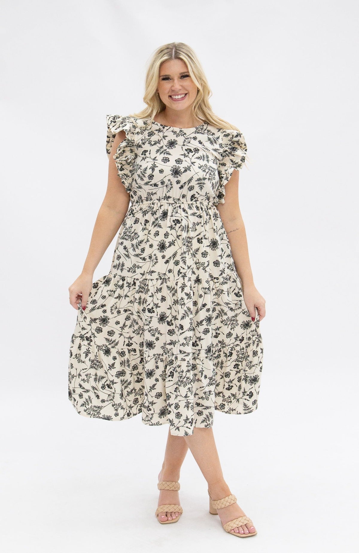 Blonde woman in a black and ivory floral tiered dress with ruffled cap sleeves, smiling against a white backdrop.