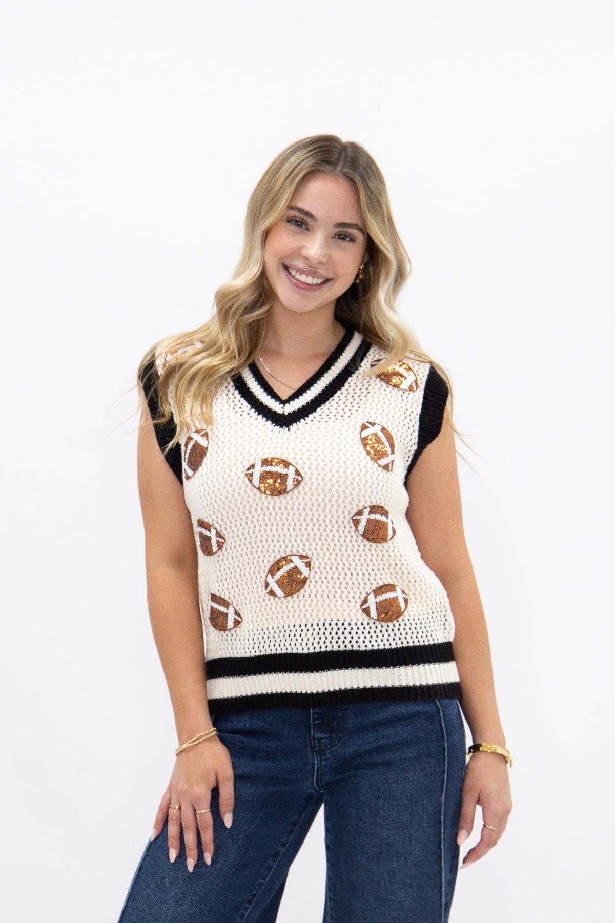 A smiling blonde woman models a white knit sweater vest with black trim and glittery brown football patches.